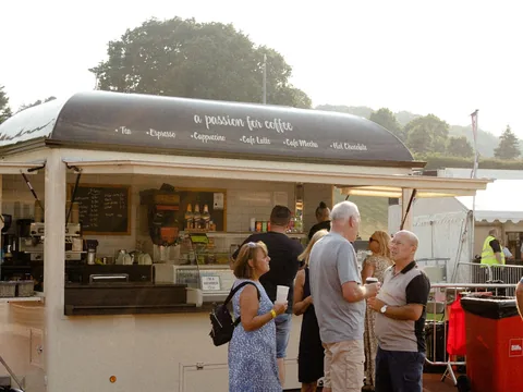 A white catering trailer on grass with a queue of people waiting at the hatch.